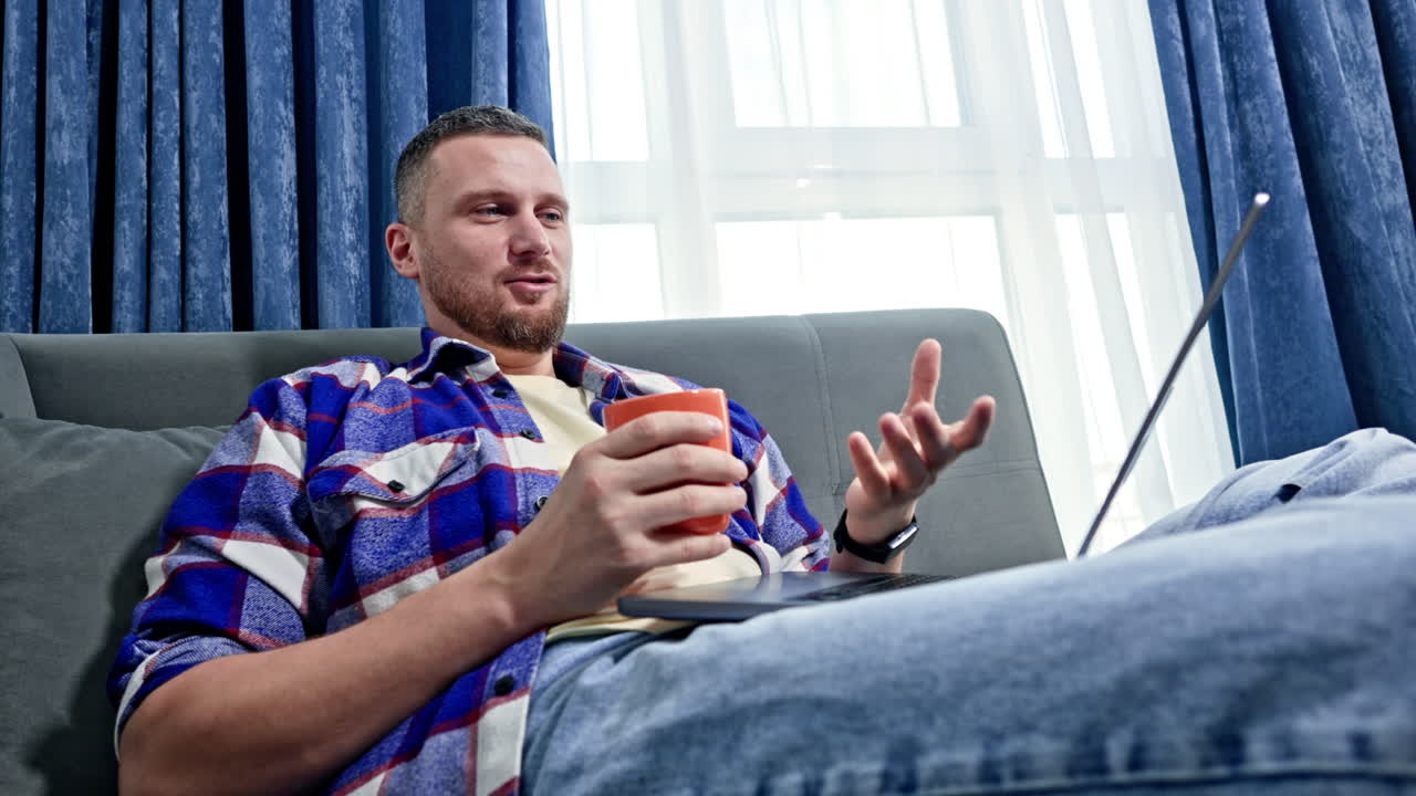 Mand holder en kop, mens han sidder på en sofa og bruger en bærbar computer i et rum med blå gardiner om dagen