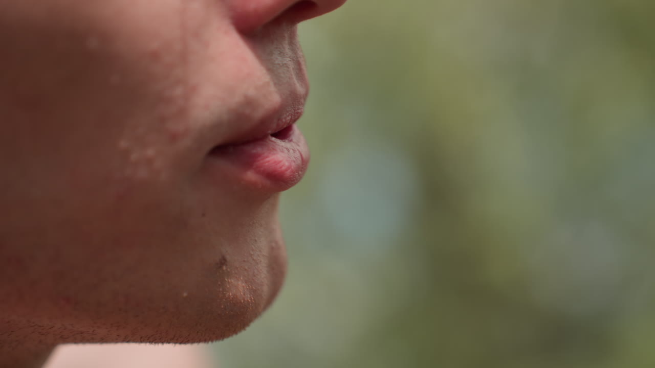 Extreme close-up of young soldier's jaw and mouth as he blows air through parted lips, revealing shaved stubble, facial texture, and slight skin bumps, against soft-focus daylight outdoor backdrop