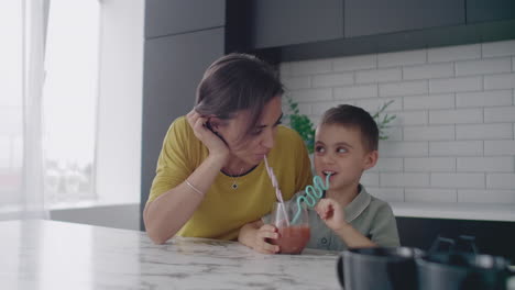 Liebevolle-Schöne-Mutter-Und-Sohn-Zusammen-Lächelnd-Trinken-Orangensaft-Aus-Einem-Glas-Durch-Eine-Röhre