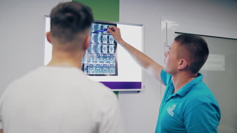 back view of a male doctor and patient examining x-ray in the hospital. specialist shows the problem areas of the back on the x-ray photograph to a young man.