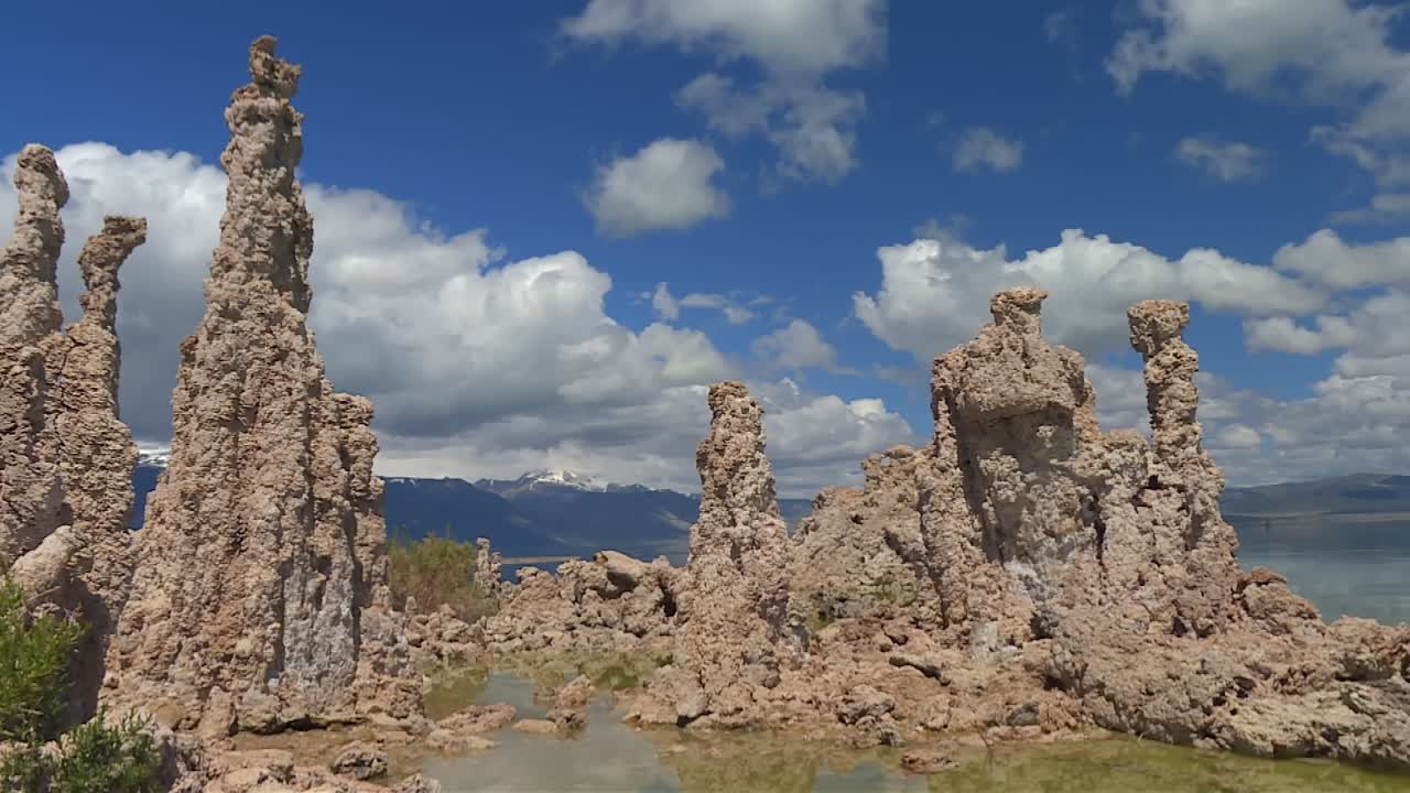 Imponentes torres de tufo ao lado de poças de água rasa sob um céu azul, com Twin Peaks visível ao fundo no Lago Mono.