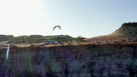 Tiro-De-Drone-Aéreo-Rotativo-Siguiendo-Un-Parapente-Volando-A-Lo-Largo-De-Un-Acantilado-Con-Grandes-Dunas-De-Arena-En-El-Fondo-Cerca-De-Pipa,-Brasil-En-Rio-Grande-Do-Norte-En-Un-Cálido-Día-De-Verano