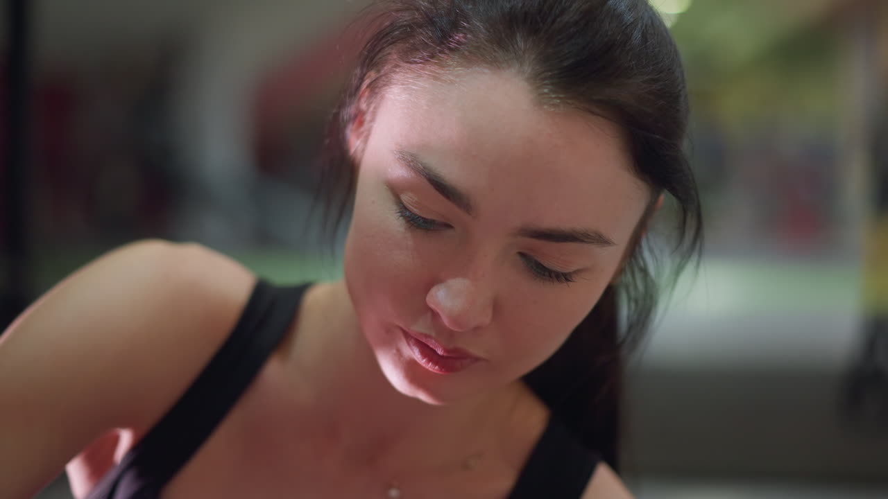 closeup gym woman sweating focused, postworkout pause as trainer reviews performance, soft gym lighting, beads of sweat glistening on skin, contemplative expression, blurred equipment background