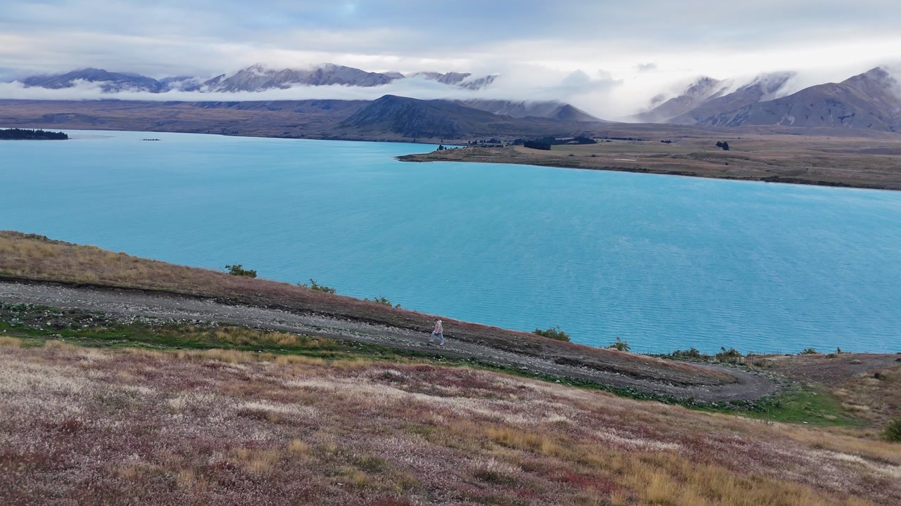 Lone tourist walks on rugged gravel road by pale blue mountain lake