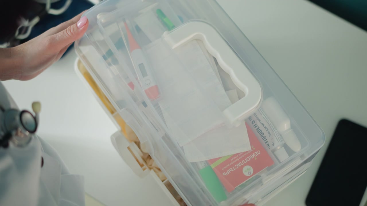 Doctor hands organizing first aid kit on desk, stethoscope visible on chest, sorting bandages and antiseptic items into clear organizer, tidy clinical atmosphere with focused professional care