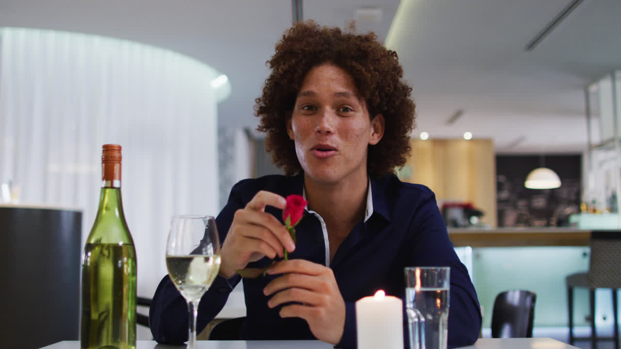 Mann av blandet etnisitet har romantisk middag på restaurant, holder rød rose og snakker til kamera