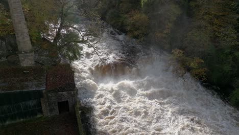 Drohnenaufnahmen,-Die-Sich-Langsam-über-Einem-Schnell-Fließenden-Fluss-Und-Einem-Wasserfall-Erheben,-Der-Von-Alten-Gebäuden-Umgeben-Ist