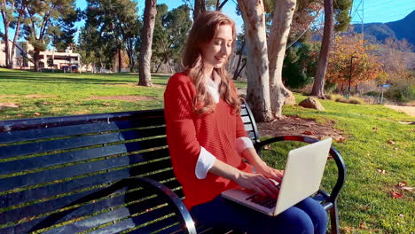 A-happy,-attractive,-young,-caucasian-woman-at-the-park,-sitting-on-a-bench,-typing-on-her-laptop,-giving-a-big-laugh