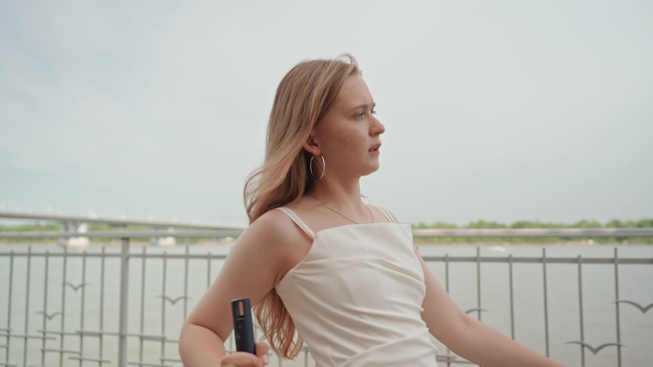Vrouw met lang haar loopt langs een promenade aan de rivier, met een voorwerp in haar hand. De scène kenmerkt zich door zacht natuurlijk licht en kalm water met groen in de verte onder een bewolkte hemel. De vrouw beweegt.