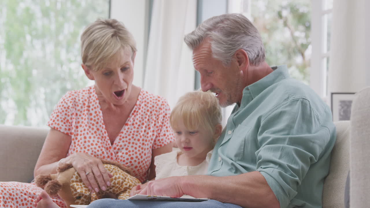 집에서 손녀와 함께 소파에 앉아 함께 책을 읽고 있는 할아버지와 할머니