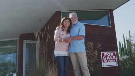 Retrato-De-Una-Pareja-Mayor-Parada-Al-Aire-Libre-Frente-A-La-Casa-Con-Un-Cartel-De-Venta-En-El-Jardín