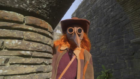 Redhead-wearing-plague-mask-stood-under-stone-archway-with-turn-to-face-camera