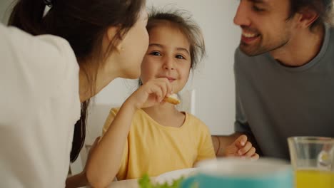 retrato de una feliz niña morena en un vestido amarillo que está desayunando y sus dos padres la besan en ambas mejillas durante el desayuno en la mesa de la familia por la mañana en un apartamento moderno