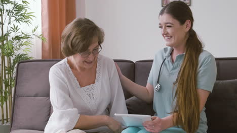 anciana y doctora sentadas en un sofá hablando y riendo mientras miran una tableta 3