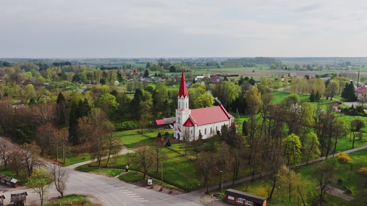 Panorama de dron revela modesta capilla blanca en medio de un paisaje rural primaveral, Rucava