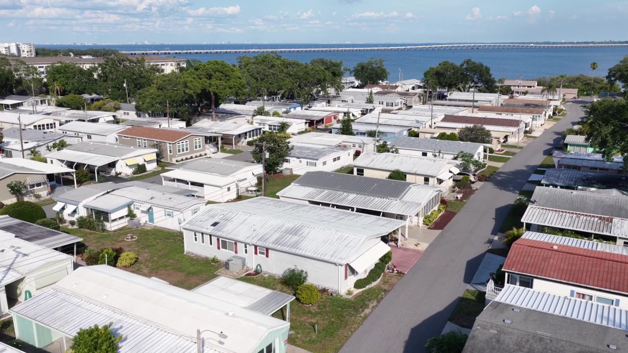 I tetti delle case mobili riempiono la fitta griglia comunitaria di Clearwater; un ponte distante si estende sulle acque scintillanti della baia sotto un cielo parzialmente nuvoloso della Florida, sopra strade alberate e ombreggiate. Ampia ripresa aerea con drone.