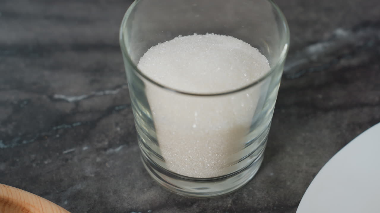 primer plano de una mano colocando un vaso transparente lleno de azúcar blanco en una mesa negra elegante, vista parcial de la bandeja de madera y los equipos de cocina en el fondo