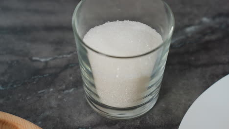 close-up of a hand placing a transparent glass filled with white sugar on a sleek black table, partial view of wooden tray and kitchen equipment in the background