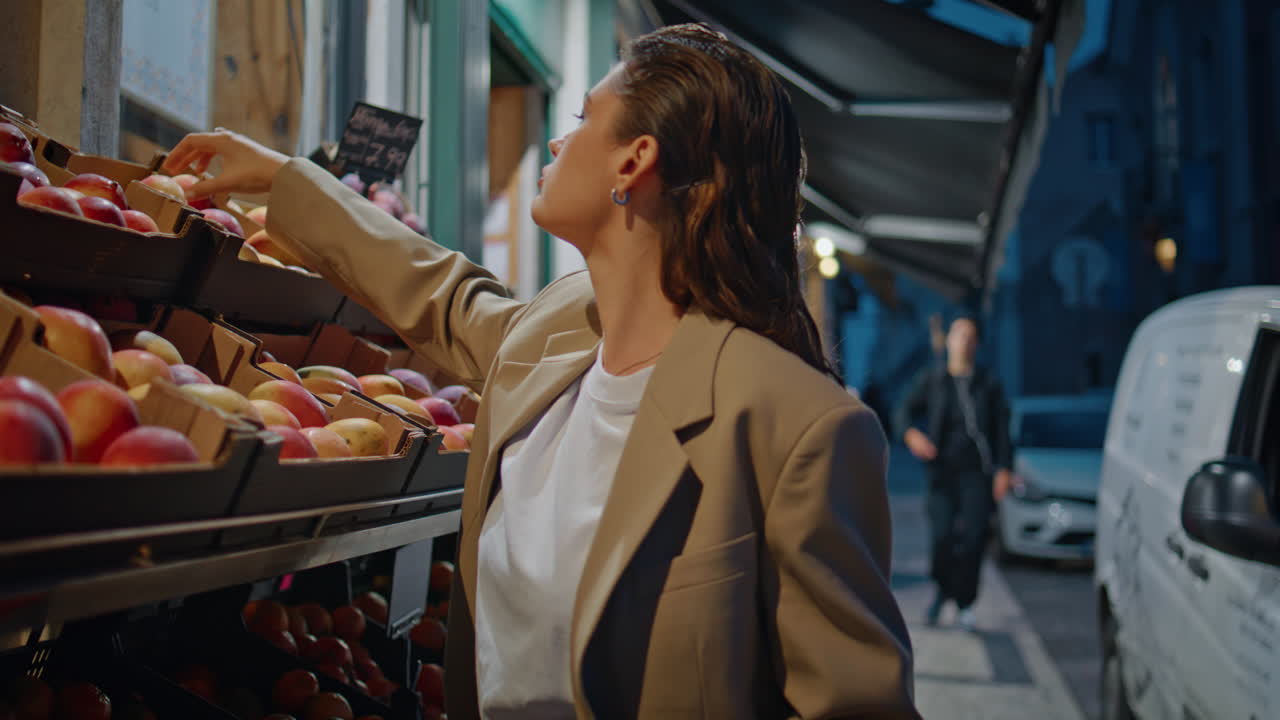 늦은 저녁에 거리의 식료품 가게에서 과일을 선택하는 젊은 여성.