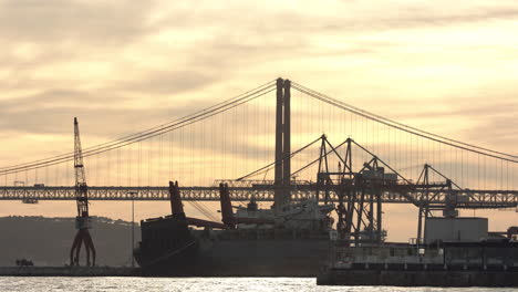 25.-April-überbrücke-Den-Fluss-Tejo-Mit-Baukränen-Und-Straßenverkehr-Im-Sonnenuntergang-Mit-Einem-Verlassenen-Frachter