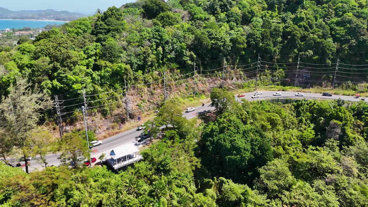 Drone footage captures vehicles navigating a winding road through lush greenery in Phuket, Thailand, under bright daylight