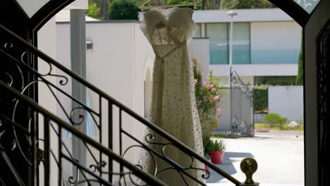 un vestido de novia blanco colgando de su percha en la entrada de una magnífica villa, vemos en primer plano las barandillas de hierro forjado de la escalera