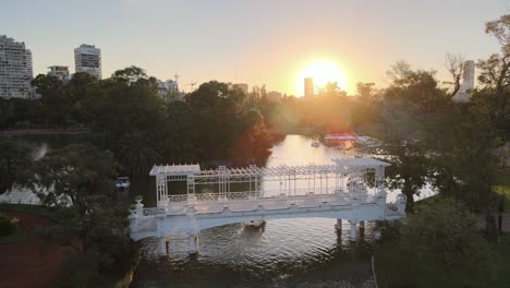 aerial rising over boats sailing under white bridge in rosedal gardens, palermo neighborhood at sunset, buenos aires