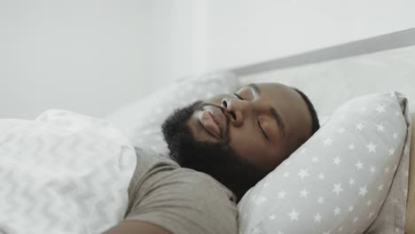 black man sleeping in bed in morning. young adult covering head with pillow.