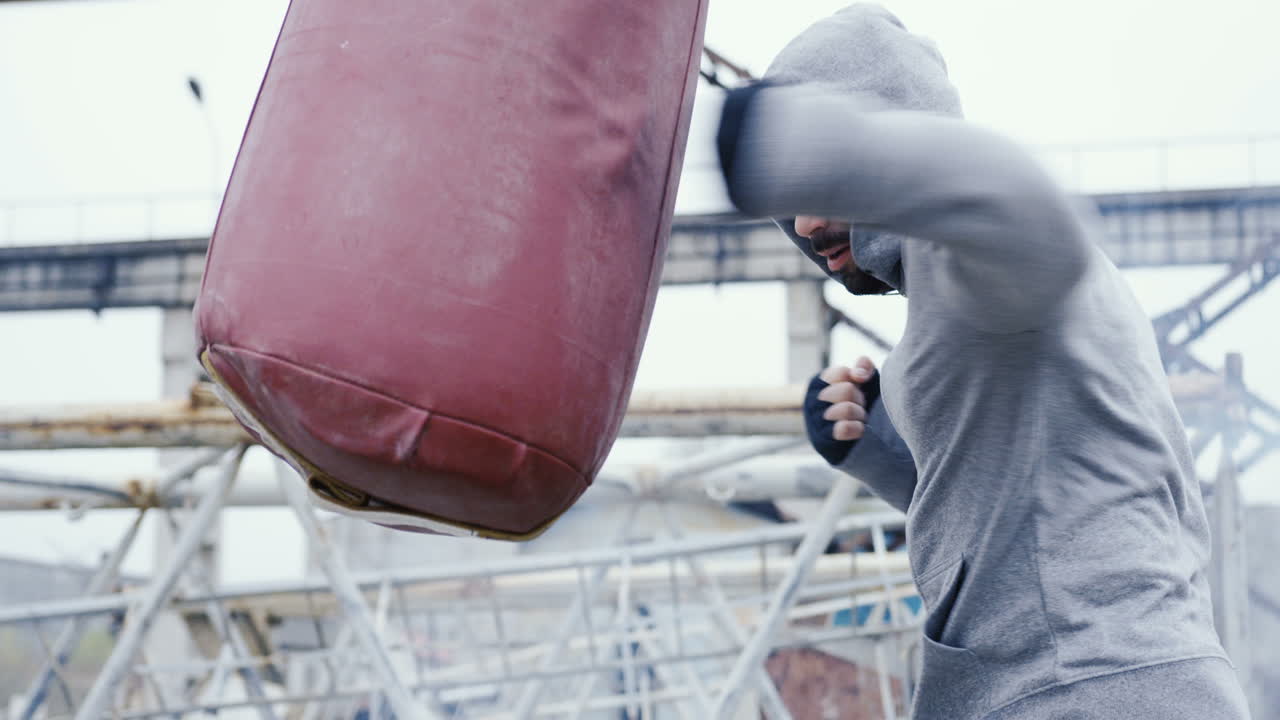 회색 후드티를 입은 백인 남자가 흐린 아침에 버려진 공장 밖에서 샌드백을 때리는 모습