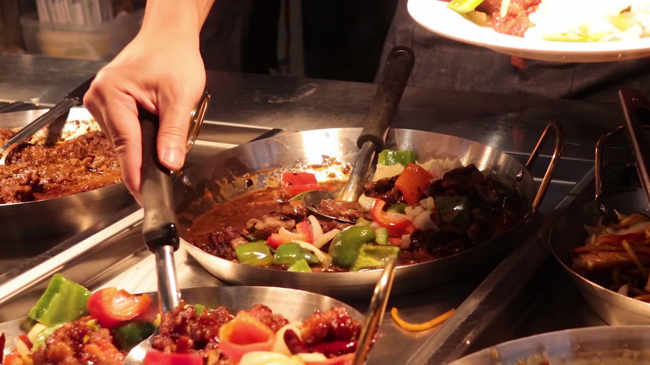Close-up of hands using tongs to serve vibrant stir-fry from a pan at a market stall.