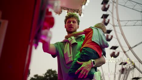 A-dad-with-curly-hair-with-stubble-in-a-green-T-shirt-holds-his-little-blond-son-in-a-red-T-shirt-who-is-shy-and-grabs-his-father's-chest-while-standing-at-the-ticket-office-in-an-amusement-park