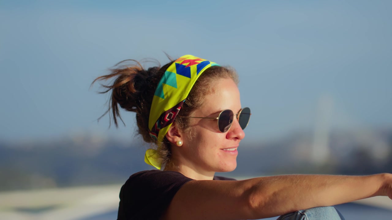 Close-up shot of a smiling and happy girl sitting by the Mondego River with Ponte Rainha Santa Isabel (Ponte Europa) in the background, Coimbra