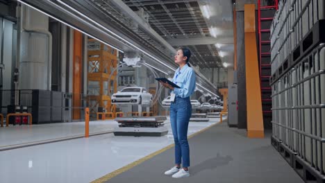 full body side view of asian female professional worker standing with tablet in ev car factory, high-tech industrial facility, she observes by looking around before she come to concentrating with tablet