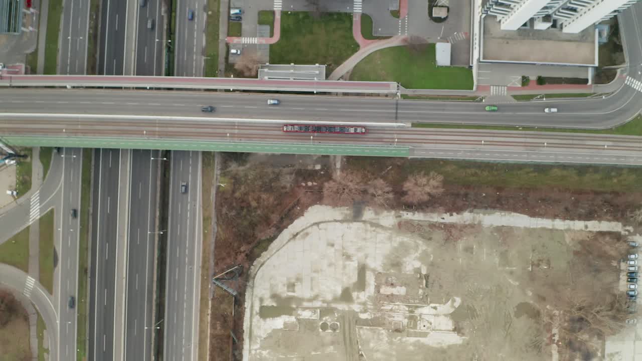 Red tram driving on a bridge crossing a highway approaching a crossroad. Aerial drone video with the camera tilted down, following the tram in traffic. Moving from right to left.
