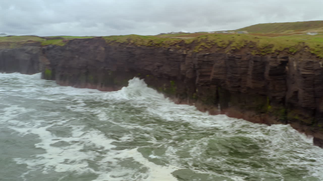 Quick aerial pan of Doolin section of the Cliffs of Moher with rough Atlantic waves under overcast skies on Ireland’s west coast