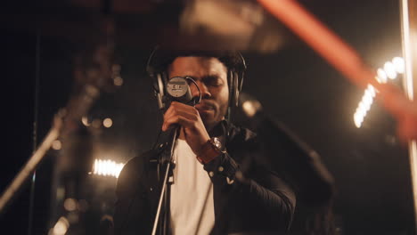 african american vocalist sings lyrical composition into microphone in sound recording studio. musician plays on drum set in soundproof room. musical band on rehearsal. concept of music production.
