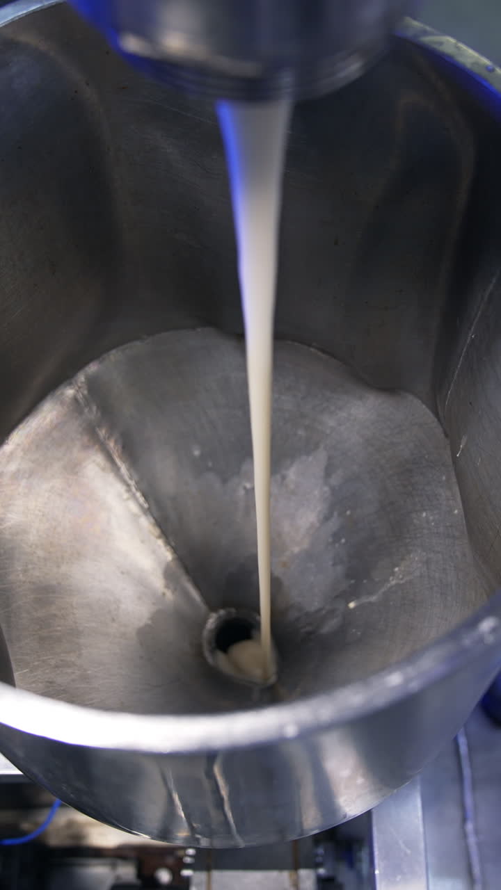 Condensed milk streaming from a tube into a metal container. Close up. Food factory automated line. View from above. Vertical video