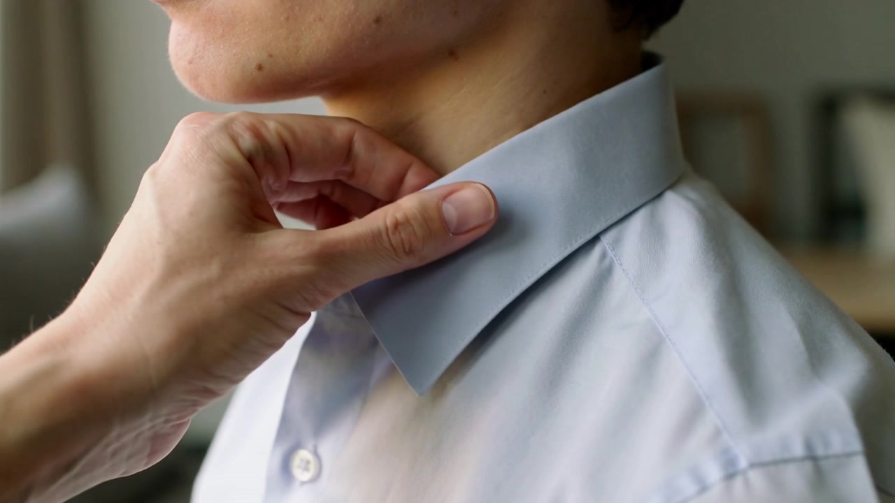 Una mano che aiuta si estende da sinistra per sistemare il colletto della camicia blu di un uomo a casa, allineandolo per un aspetto ordinato.