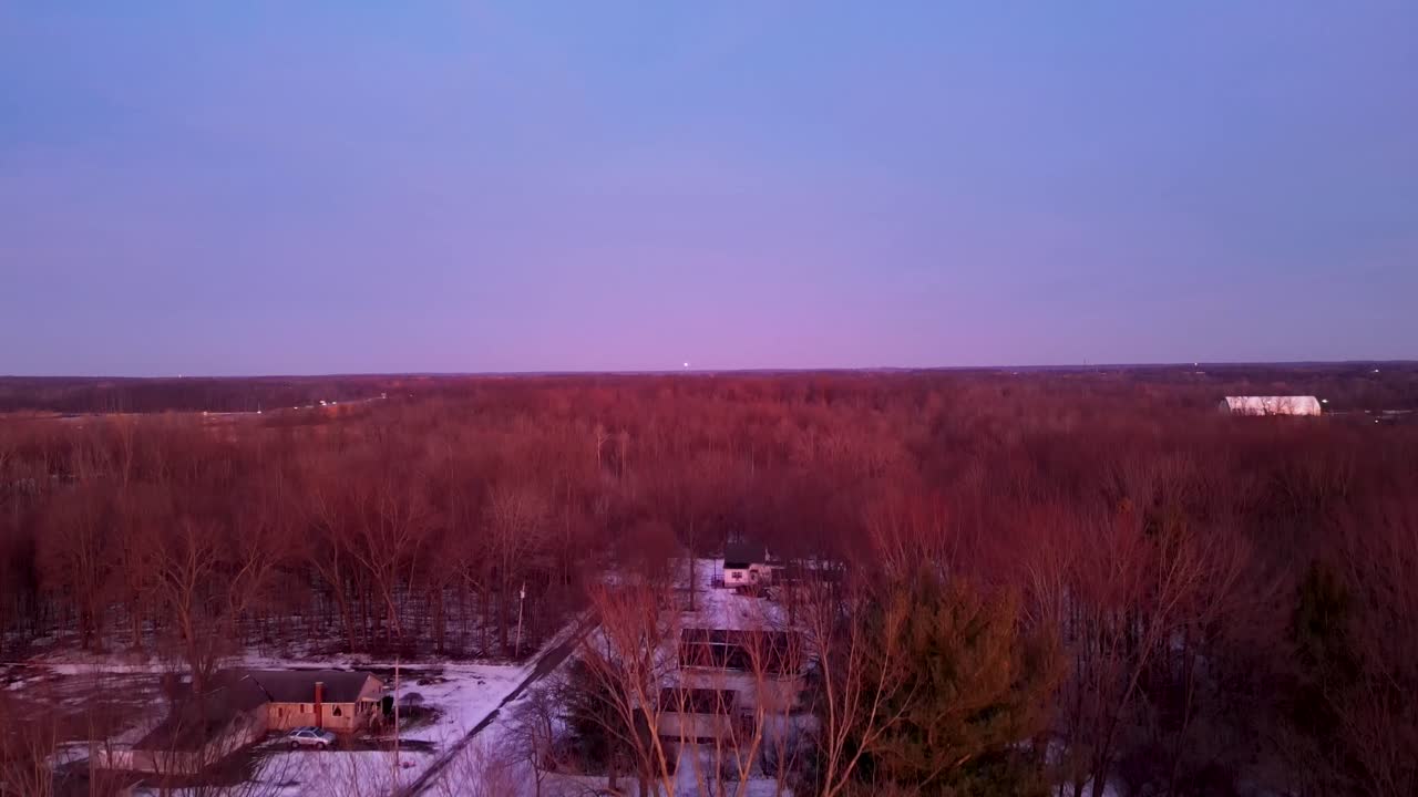 Capture of a winter sunset casting warm hues on bare trees and snow-covered rooftops, illustrating the contrast between the cold season and the warm glow of the setting sun
