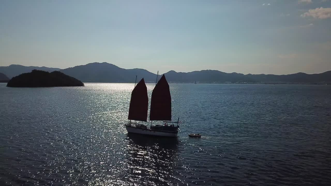 Drone shot of a moving sailing boat crossing sun flare on the water.