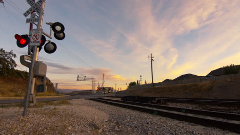 at a railway stop on a road a train has just passed and the gate is rising while the lights are flashing and cars begin to cross the tracks