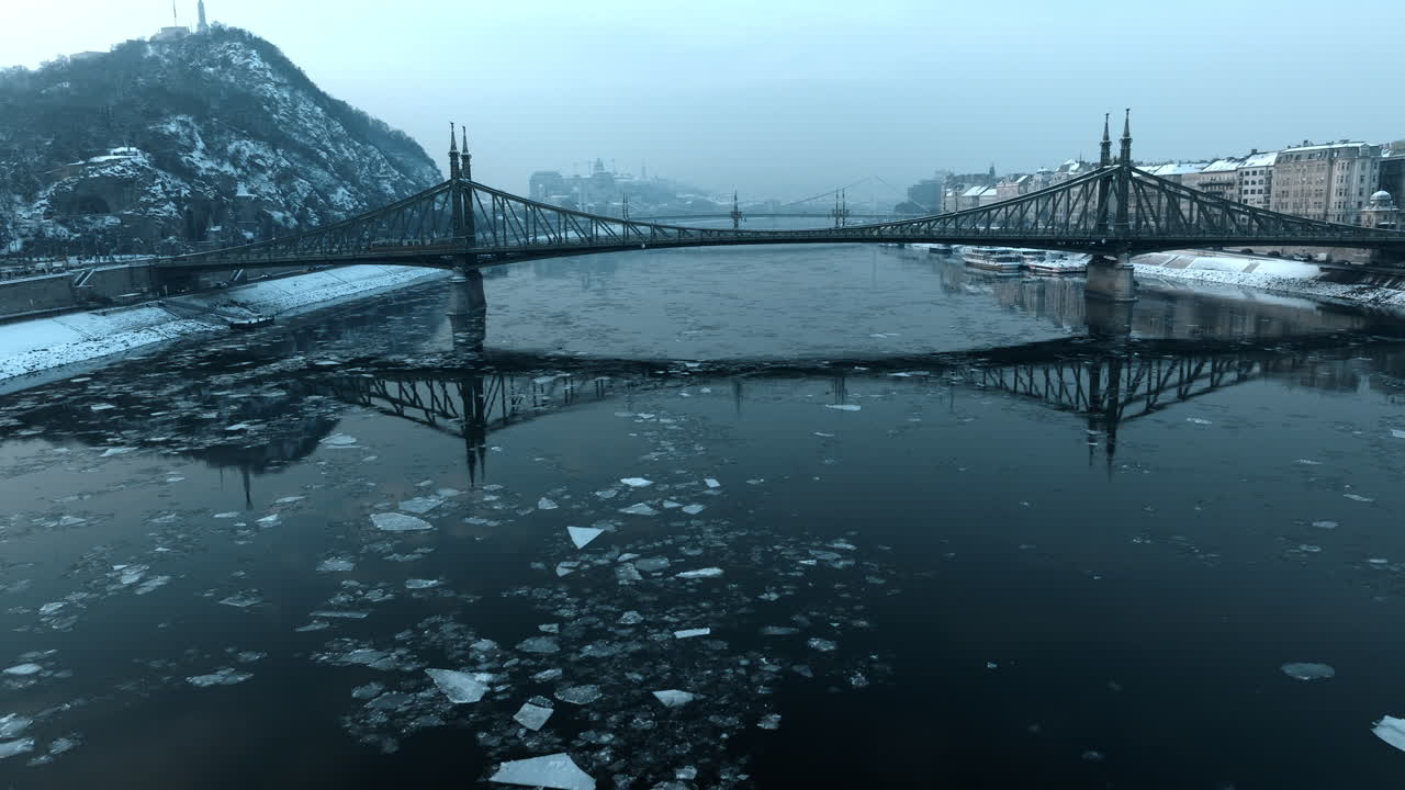 Winterse luchtfoto van een passagiersboot die door de met ijs bedekte Donau vaart onder de Vrijheidsbrug in Boedapest, met bevroren water, sneeuwval en een koude, Europese stadsatmosfeer