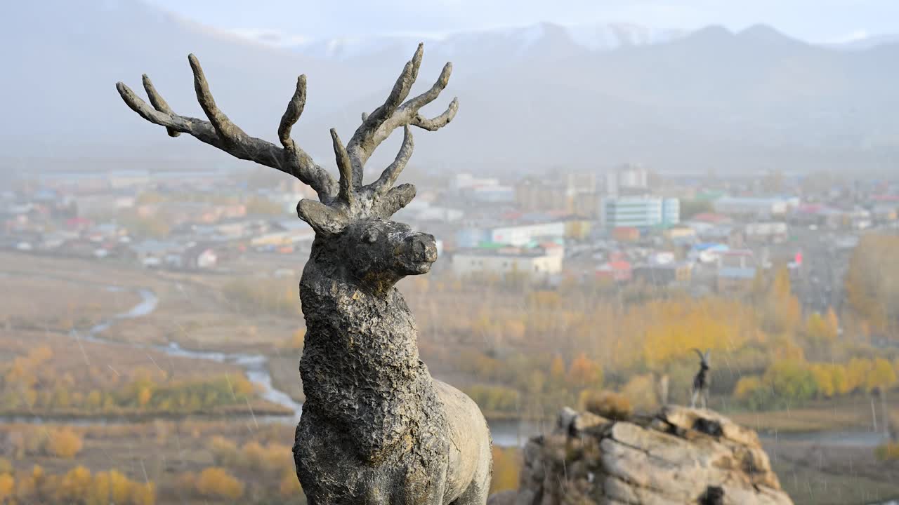 A close-up of a majestic stone stag sculpture with large antlers overlooking the town of Uliastai, Mongolia. Light snow falls on the scenic autumn valley in the background