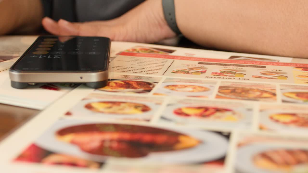 Person points at illustrated menu, deciding food order at restaurant table with smartphone and calculator