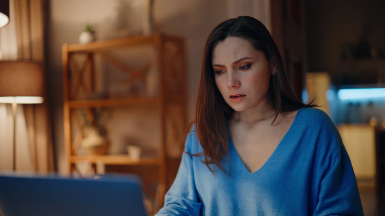 Concerned brunette reading email at laptop in dark home closeup. Desperate woman