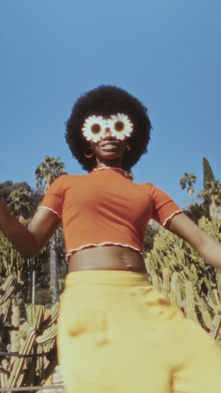 Giovane donna di colore con capelli afro e occhiali da sole a forma di margherita che balla all'aperto