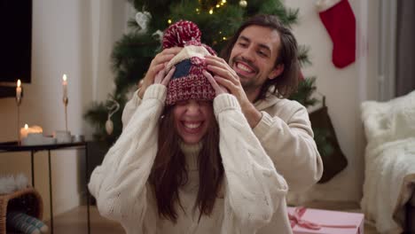 Portrait-of-a-young-brunette-guy-puts-a-woolen-New-Year's-hat-on-his-girlfriend's-head-in-a-White-sweater-in-a-room-with-a-New-Year's-tree-and-a-cozy-atmosphere-in-a-winter-evening