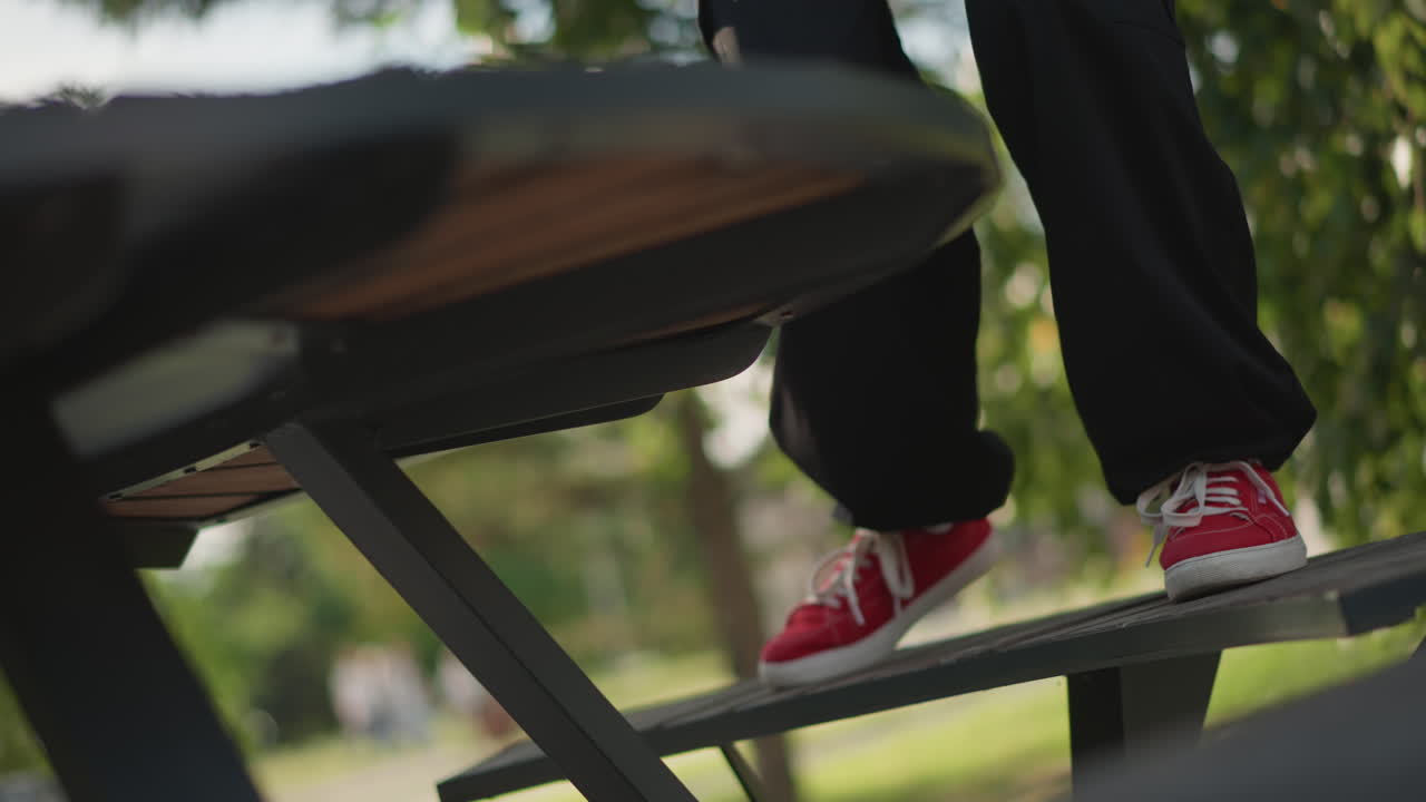Røde sneakers på bænk, gadedanser der øver indviklet fodarbejde under løvfyldt parkhimmel, sorte bukser, metalbordstel, dynamisk stepping og balance, urban øvestemning, blødt sollys