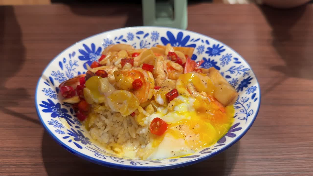 A hand uses a fork and spoon to scoop shrimp, rice, egg, and chili from a decorative bowl under bright indoor lighting, emphasizing vibrant colors and textures