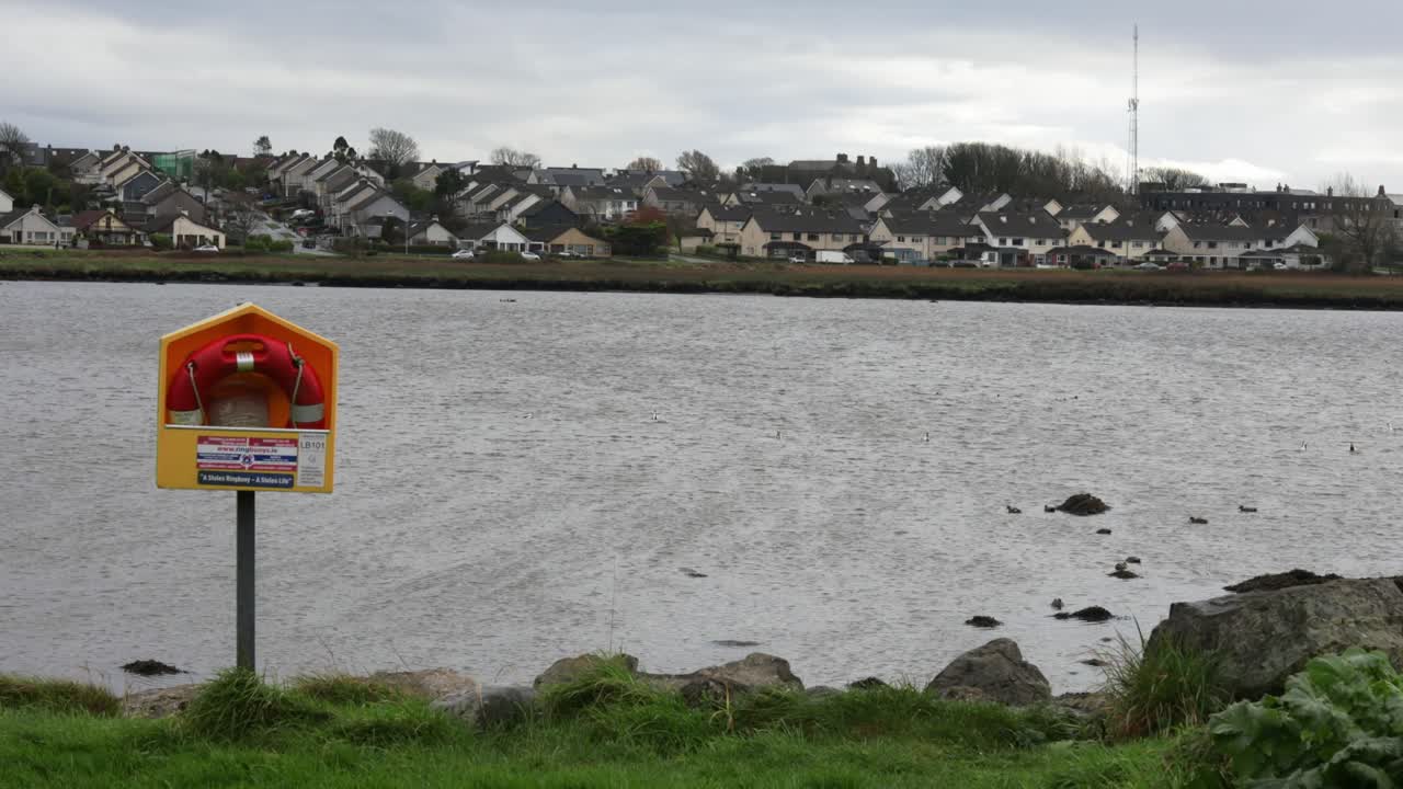 Lifebuoy station positioned along the grassy banks of Lough Atalia, with the Galway city suburbs in the background